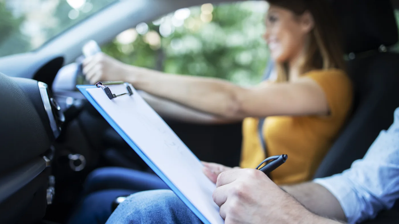 A driving instructor making notes on a clipboard beside a learner at the wheel