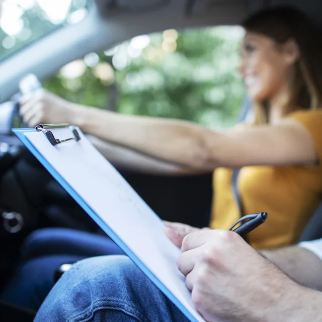 A driving instructor making notes on a clipboard beside a learner at the wheel